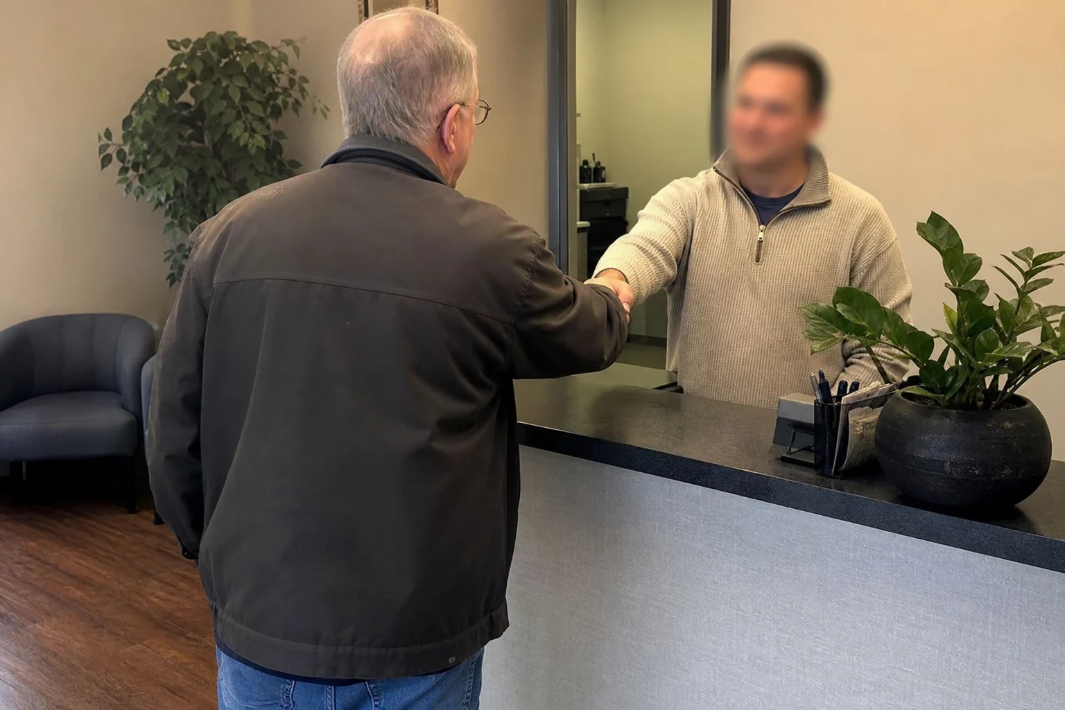Older man in a dark jacket shakes hands with an agent across a reception counter in a small office, with potted plants and a gray waiting chair nearby