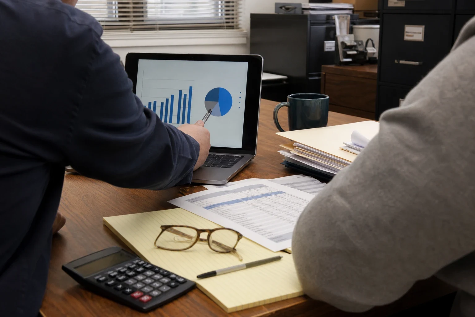 Two people at a wooden office desk reviewing bar and pie charts on a laptop, with a spreadsheet printout, legal pad, glasses, calculator and file folders nearby